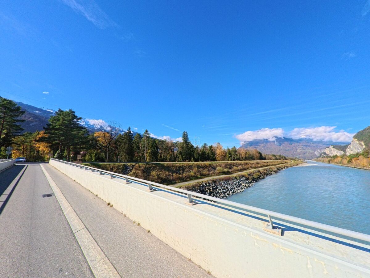 Von einer Brücke aus verläuft der Blick entlang eines breiten Flussufers mit begleitendem Weg und herbstlichem Wald nahe Fläsch (7306, Kanton Graubünden) unter klarem Himmel.