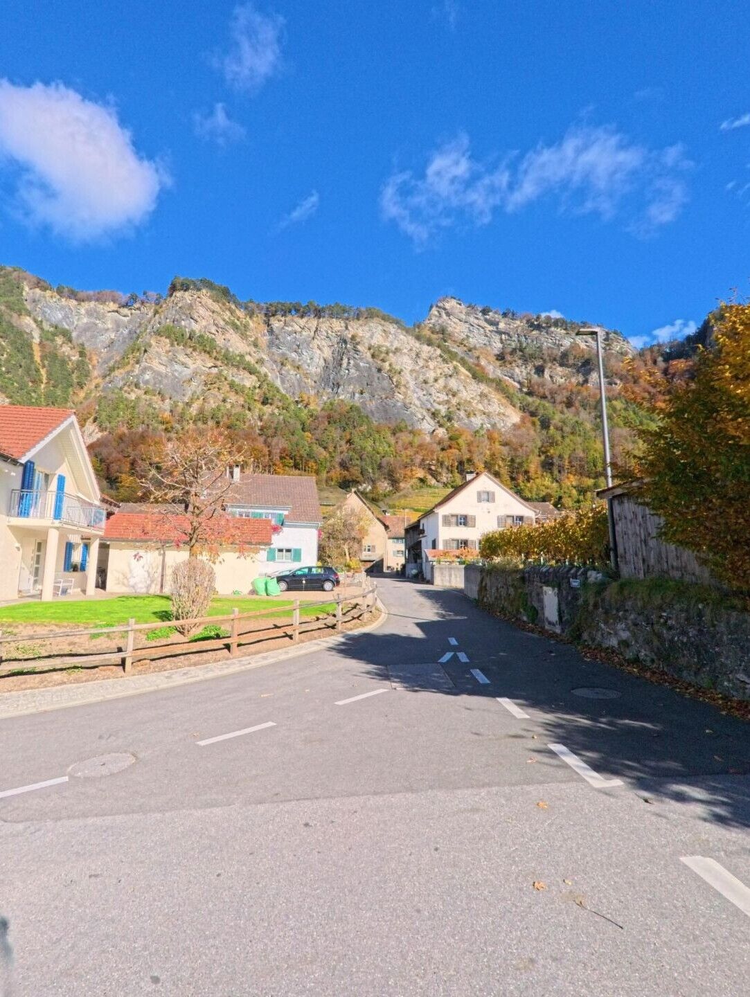 Eine leicht geschwungene Dorfstrasse führt zwischen Wohnhäusern hindurch, während sich im Hintergrund die steile Felswand bei Fläsch (7306, Kanton Graubünden) erhebt.