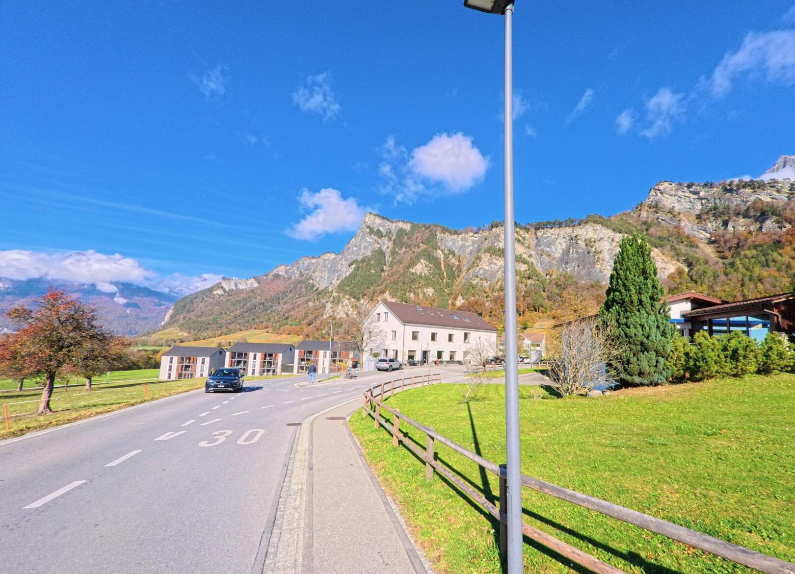 Entlang einer Tempo-30-Strasse mit angrenzenden Wiesen und Wohnhäusern zeigt sich die Bergkulisse bei Fläsch (7306, Kanton Graubünden) unter klarem Himmel.