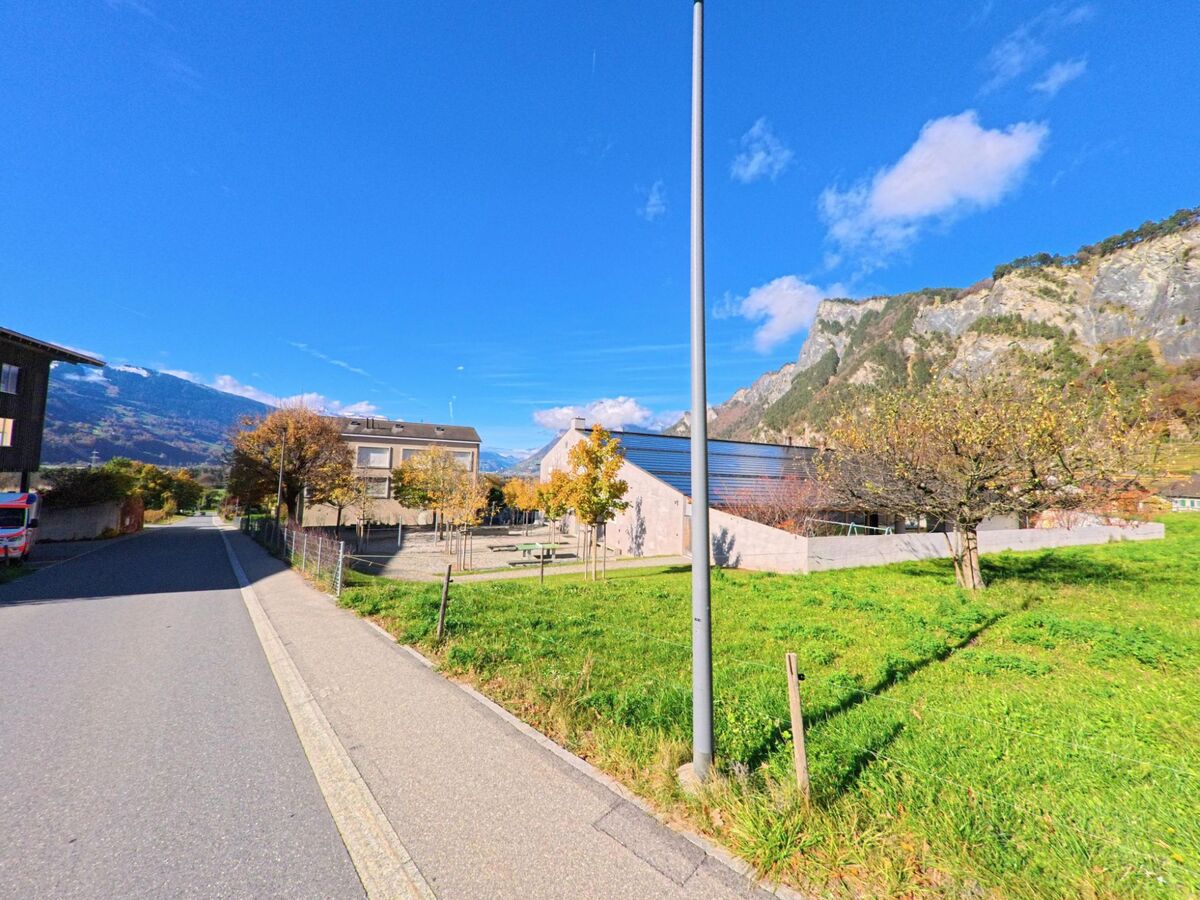Entlang einer ruhigen Strasse mit Grünflächen und öffentlichen Gebäuden zeigt sich ein Wohnbereich von Fläsch (7306, Kanton Graubünden) vor steiler Felskulisse im Herbst.