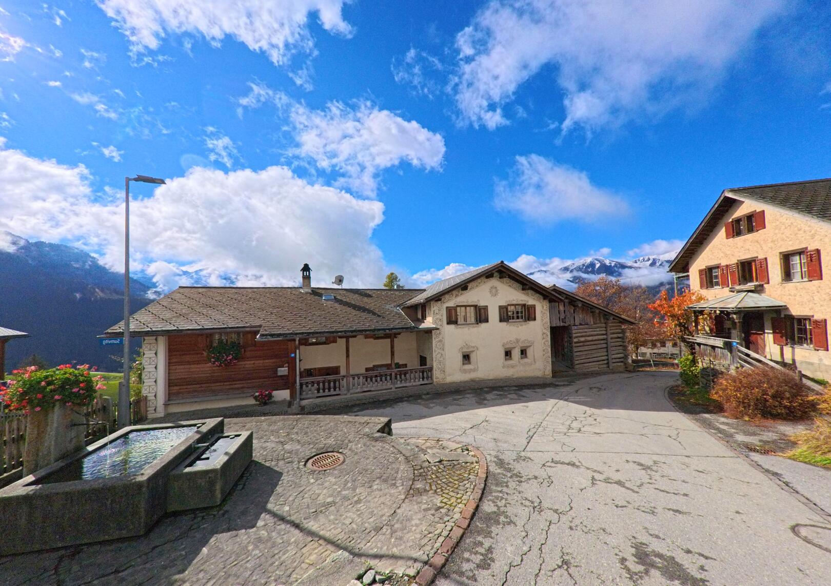 Auf einem kleinen Dorfplatz mit Brunnen und traditionellen Wohnhäusern zeigt sich das historische Ortsbild von Flerden (7426, Kanton Graubünden) unter wechselnd bewölktem Himmel.