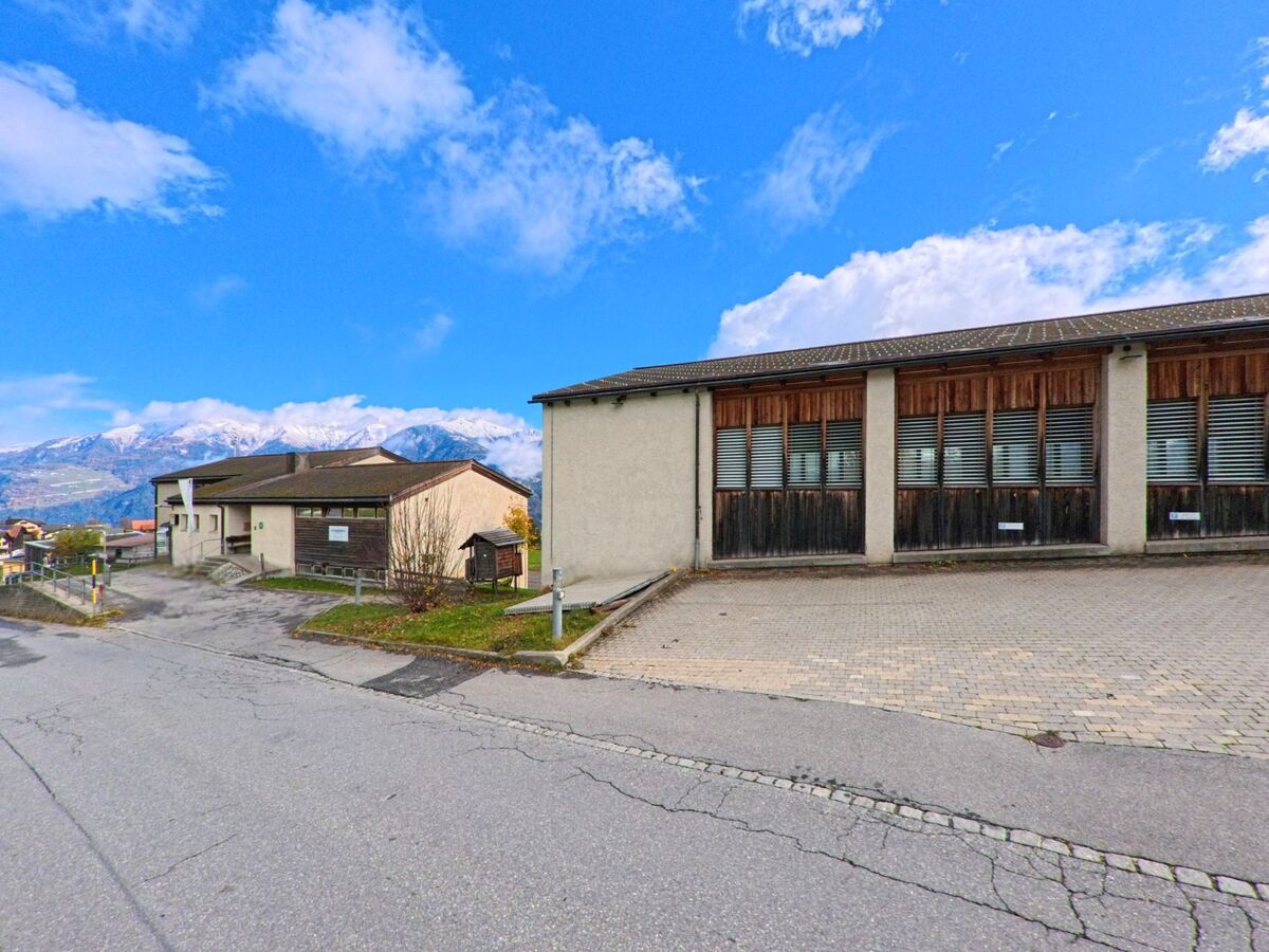 Vor funktionalen Gemeindebauten mit Vorplatz öffnet sich der Blick auf die umliegende Berglandschaft von Flerden (7426, Kanton Graubünden) unter aufgelockerter Bewölkung.