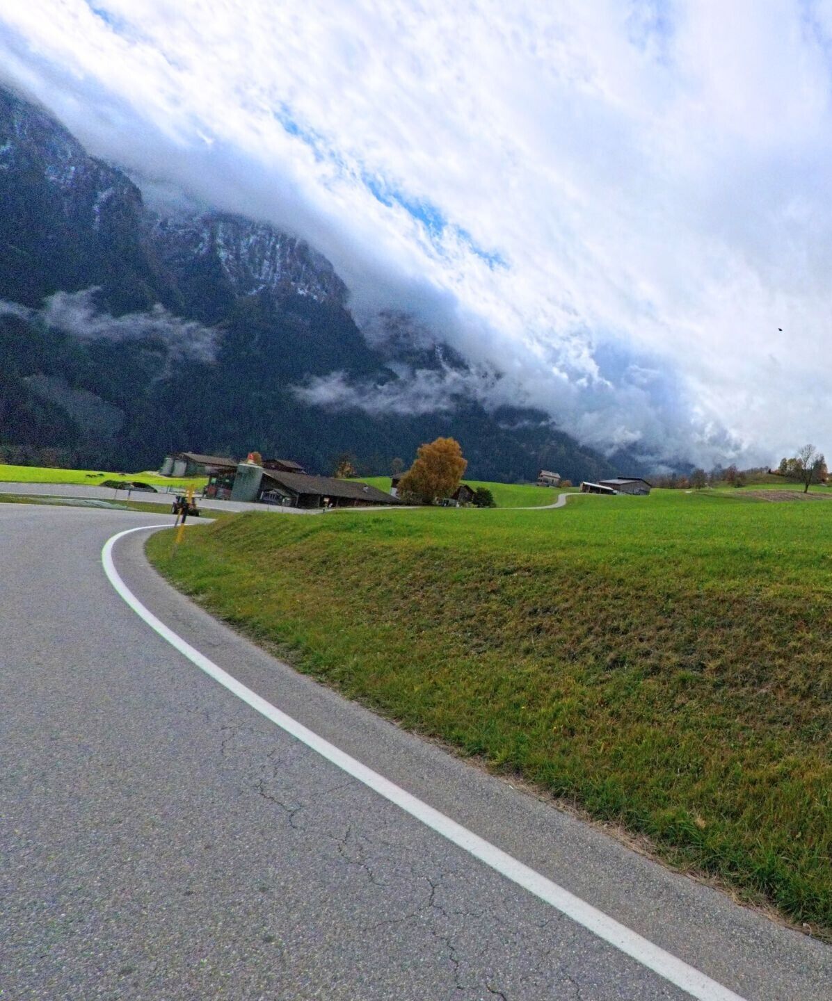Eine geschwungene Landstrasse führt durch offene Wiesen, während tief hängende Wolken die Bergflanken bei Flerden (7426, Kanton Graubünden) teilweise umhüllen.