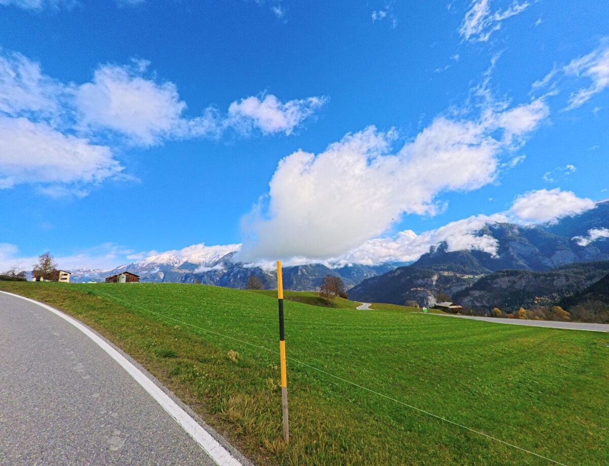 Eine leicht geschwungene Strasse verläuft entlang weiter Wiesen, während Wolken über der Berglandschaft von Flerden (7426, Kanton Graubünden) ziehen.