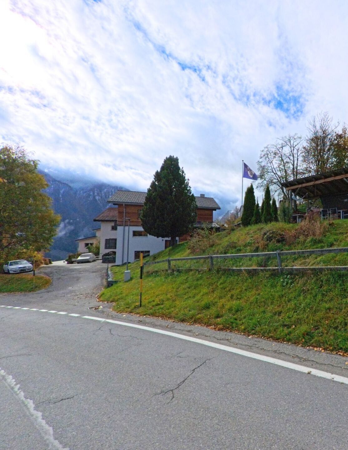 An einer leicht ansteigenden Strassenkurve stehen Wohnhäuser am Hang, während Fahne und Bäume das Ortsbild von Flerden (7426, Kanton Graubünden) prägen.