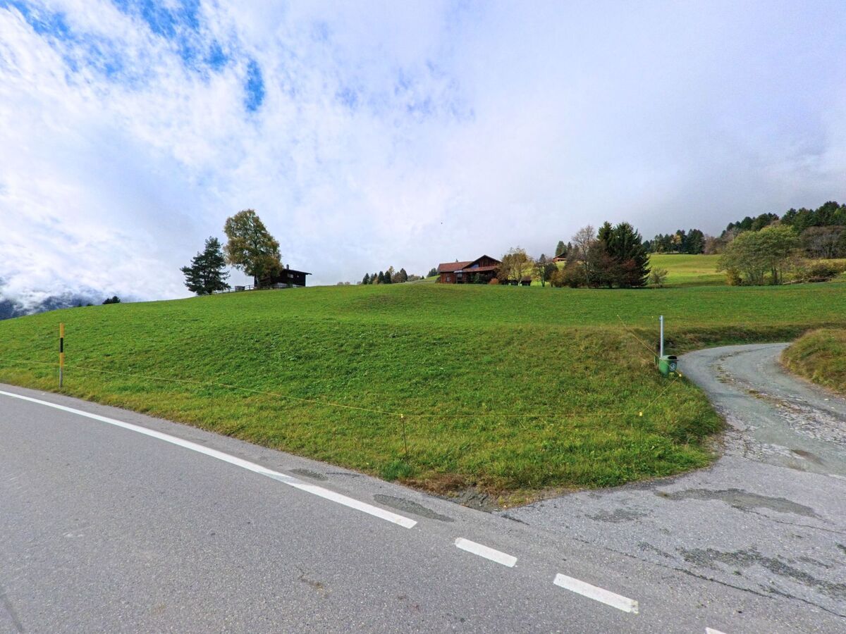Eine weite, leicht ansteigende Wiesenfläche mit einzelnen Gebäuden prägt die ruhige Landschaft am Rand von Flerden (7426, Kanton Graubünden) unter wechselnd bewölktem Himmel.