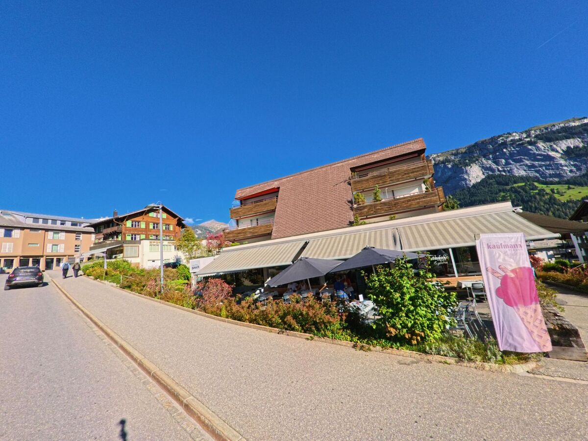 An einer leicht ansteigenden Strasse in Flims (7018, Kanton Graubuenden) befinden sich mehrere Wohnhaeuser und ein Cafe mit Sonnenterrasse vor der Bergkulisse.