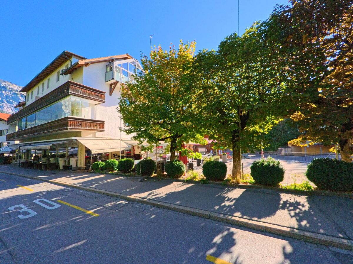 Ein Gebaeude mit grossem Balkon und verglaster Fassade steht in Flims (7018, Kanton Graubuenden) an einer von Baeumen gesaeumten Strasse bei Sonnenschein.