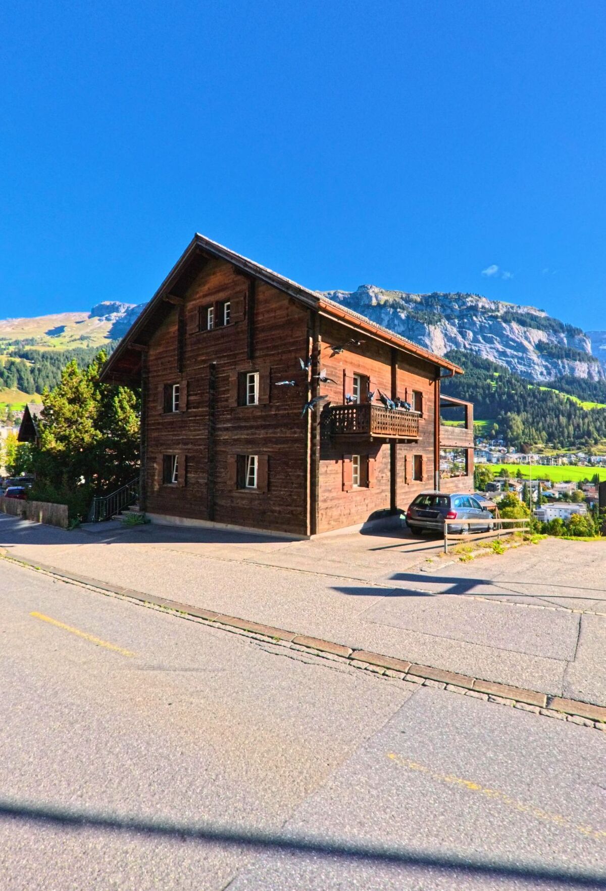 Ein traditionelles Holzhaus in Flims (7018, Kanton Graubuenden) steht an einer leicht ansteigenden Strasse mit Blick auf die umliegenden Berge.