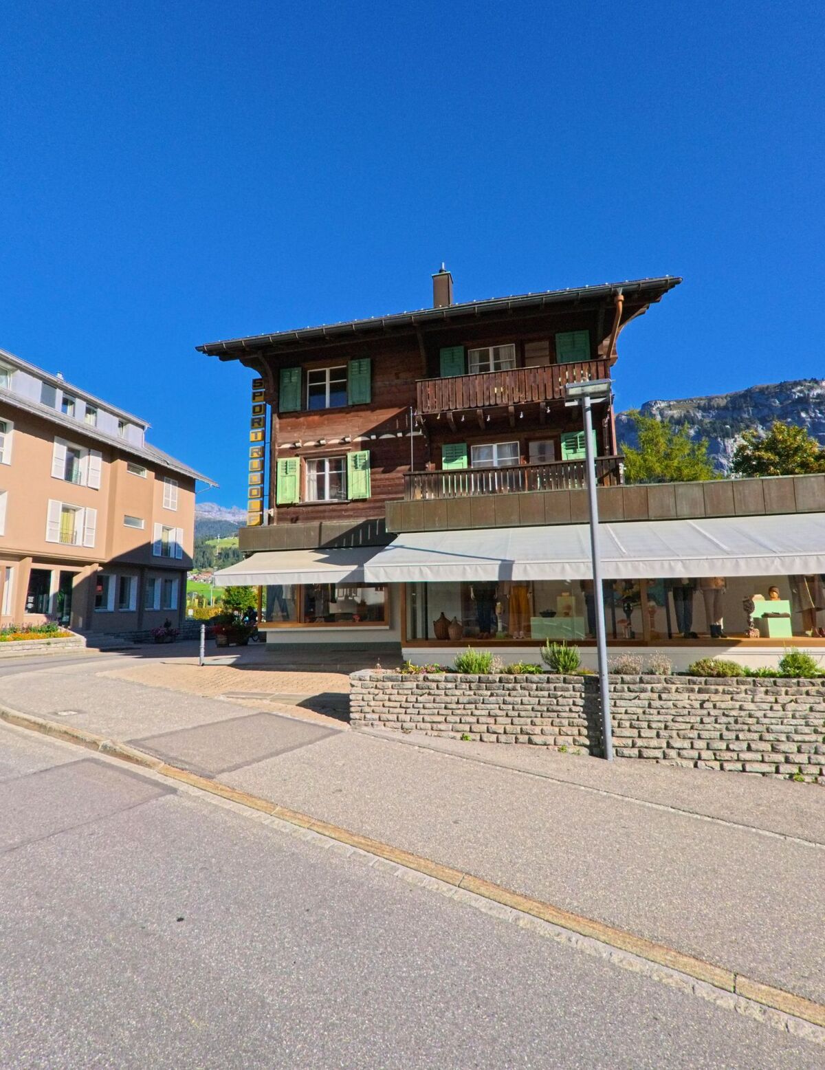 In Flims (7018, Kanton Graubuenden) steht ein traditionelles Holzhaus mit gruenen Fensterlaeden und Ladenzeile im Erdgeschoss an einer sonnigen Strasse.