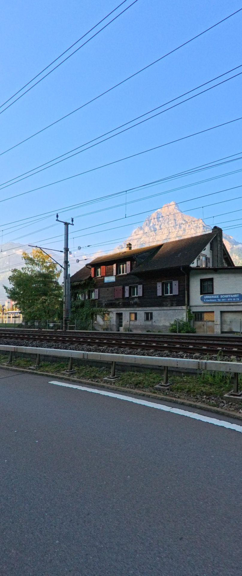 Entlang der Bahnlinie in Flüelen (6454, Kanton Uri) verläuft eine Strasse mit Blick auf ein historisches Gebäude und die markanten Berggipfel im Hintergrund.