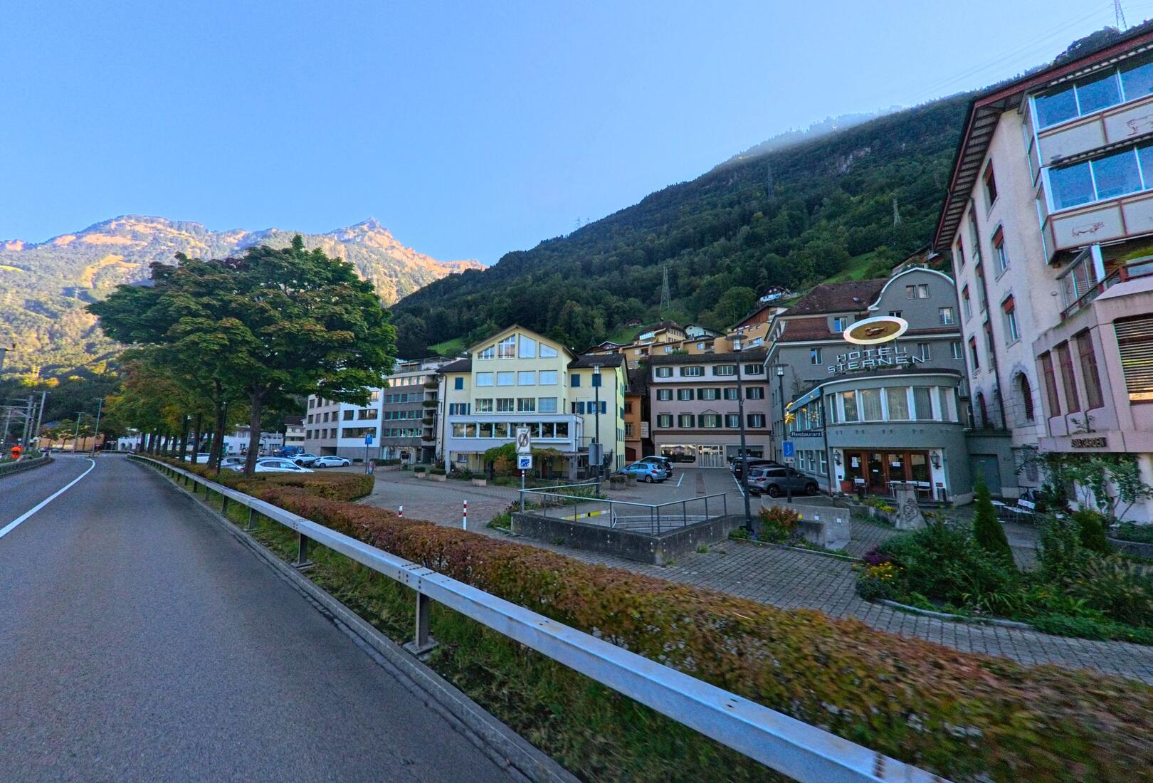 Entlang einer ruhigen Strasse in Flüelen (6454, Kanton Uri) zeigt sich ein Dorfbereich mit Hotels, Wohnhäusern und bewaldeten Berghängen im Hintergrund.