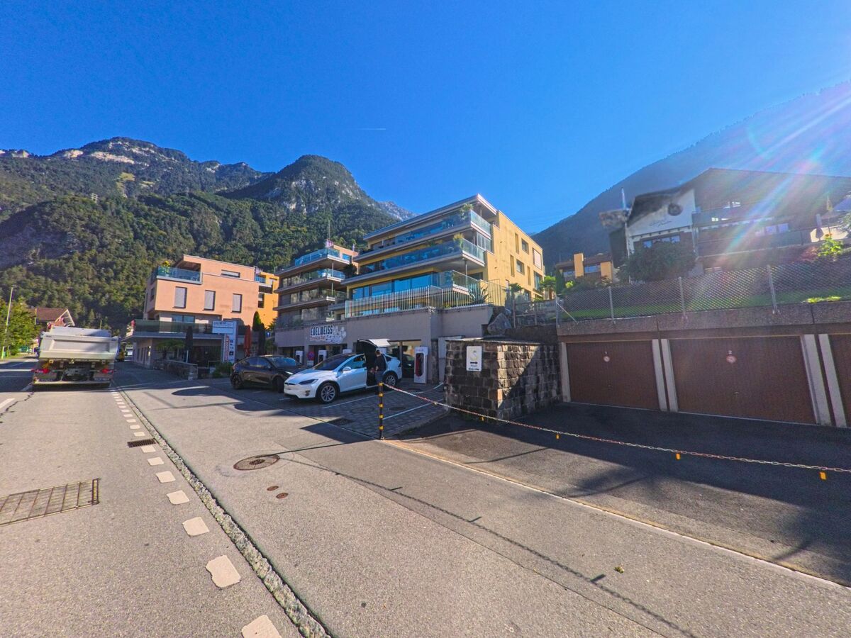 Entlang einer Strasse in Flüelen (6454, Kanton Uri) reihen sich moderne Wohn- und Geschäftsgebäude mit Parkplätzen, eingerahmt von steilen, bewaldeten Berghängen.