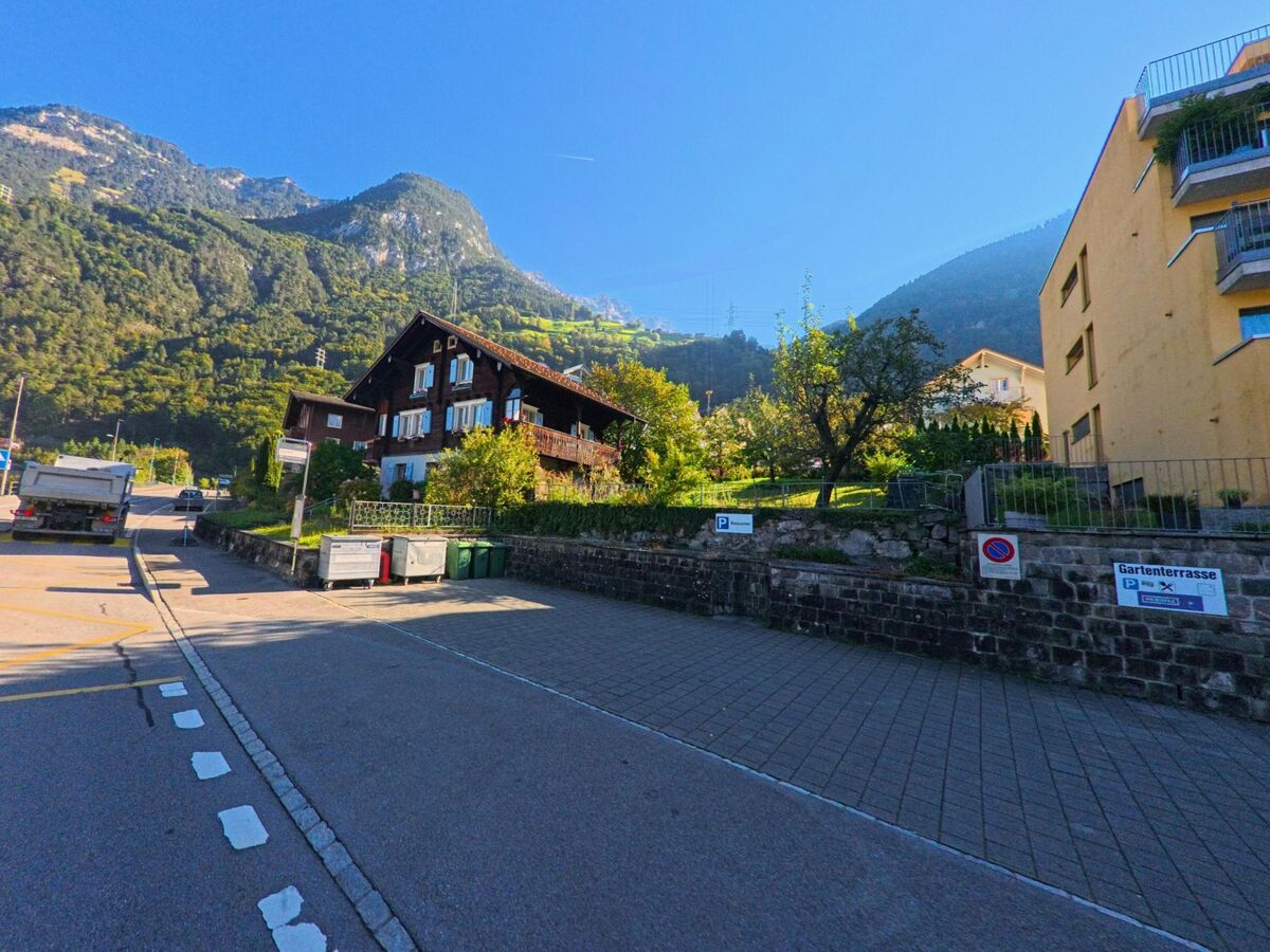 Am Strassenrand von Flüelen (6454, Kanton Uri) liegen Wohnhäuser mit Gärten, während sich dahinter die bewaldeten Berghänge unter klarem Himmel erheben.