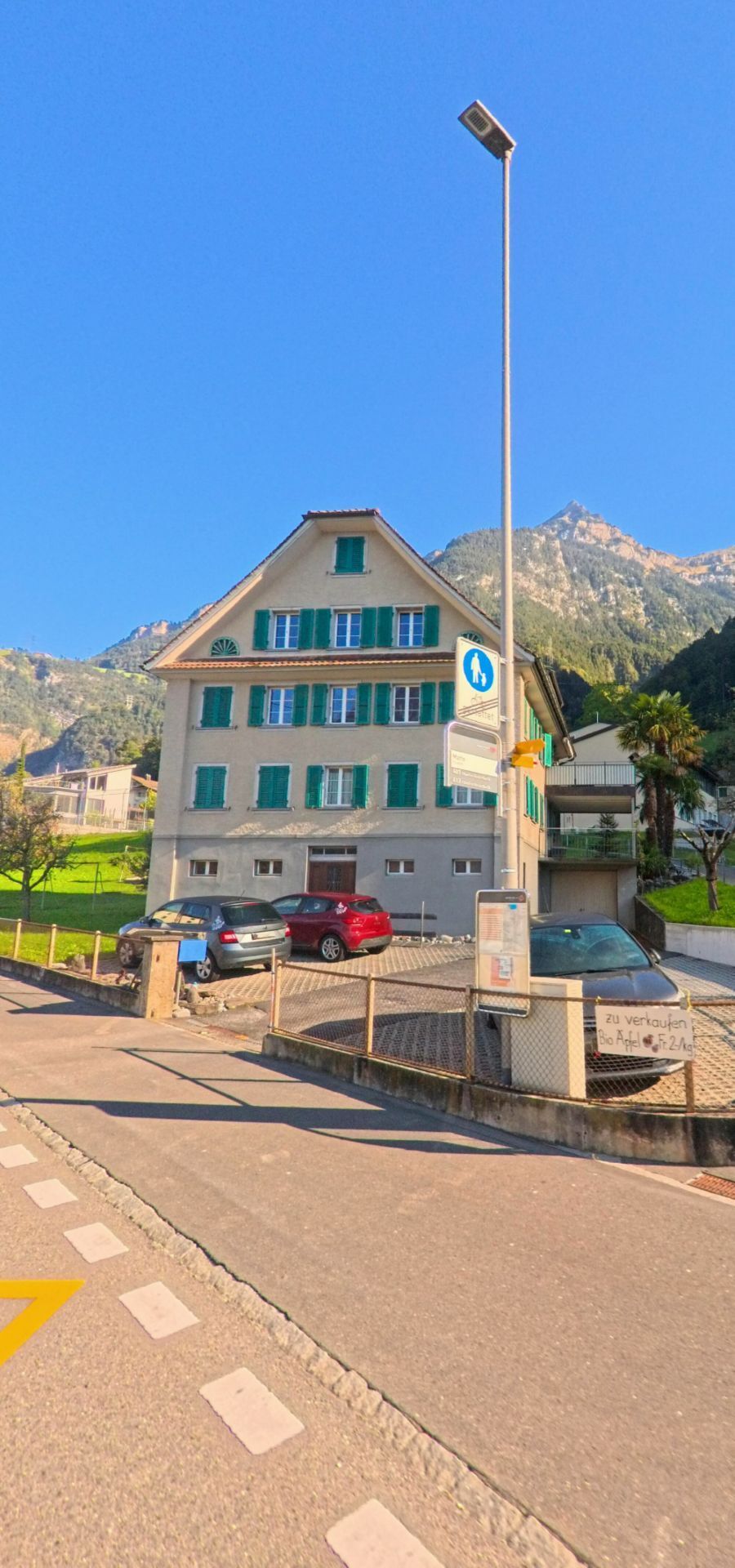 An einer Strassenkreuzung in Flüelen (6454, Kanton Uri) steht ein Wohnhaus mit grünen Fensterläden, davor Parkplätze und Verkehrszeichen vor der alpinen Bergkulisse.