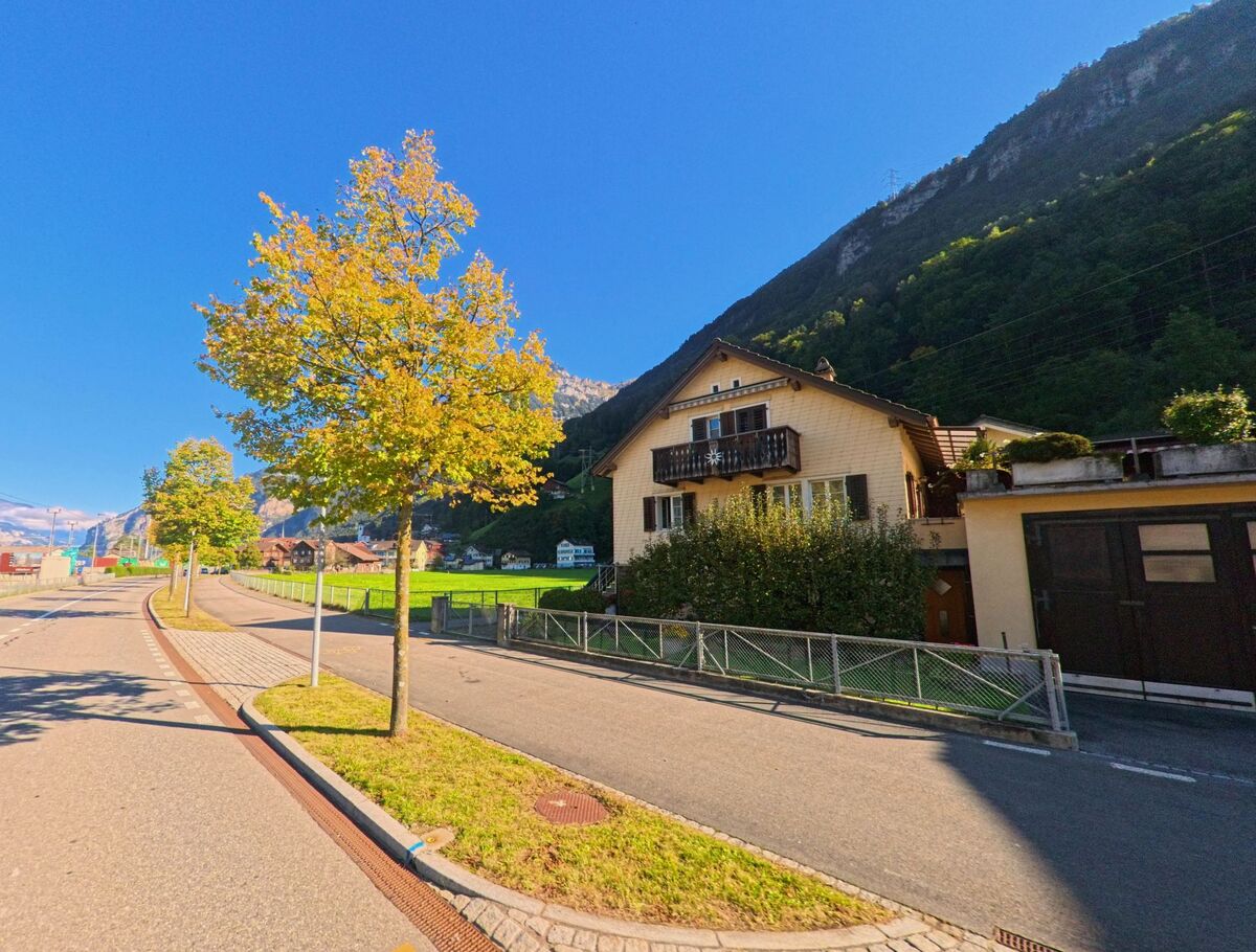 Neben einer Wohnstrasse in Flüelen (6454, Kanton Uri) liegen Einfamilienhäuser und offene Grünflächen, während herbstlich gefärbte Bäume den Blick zum Berghang begleiten.