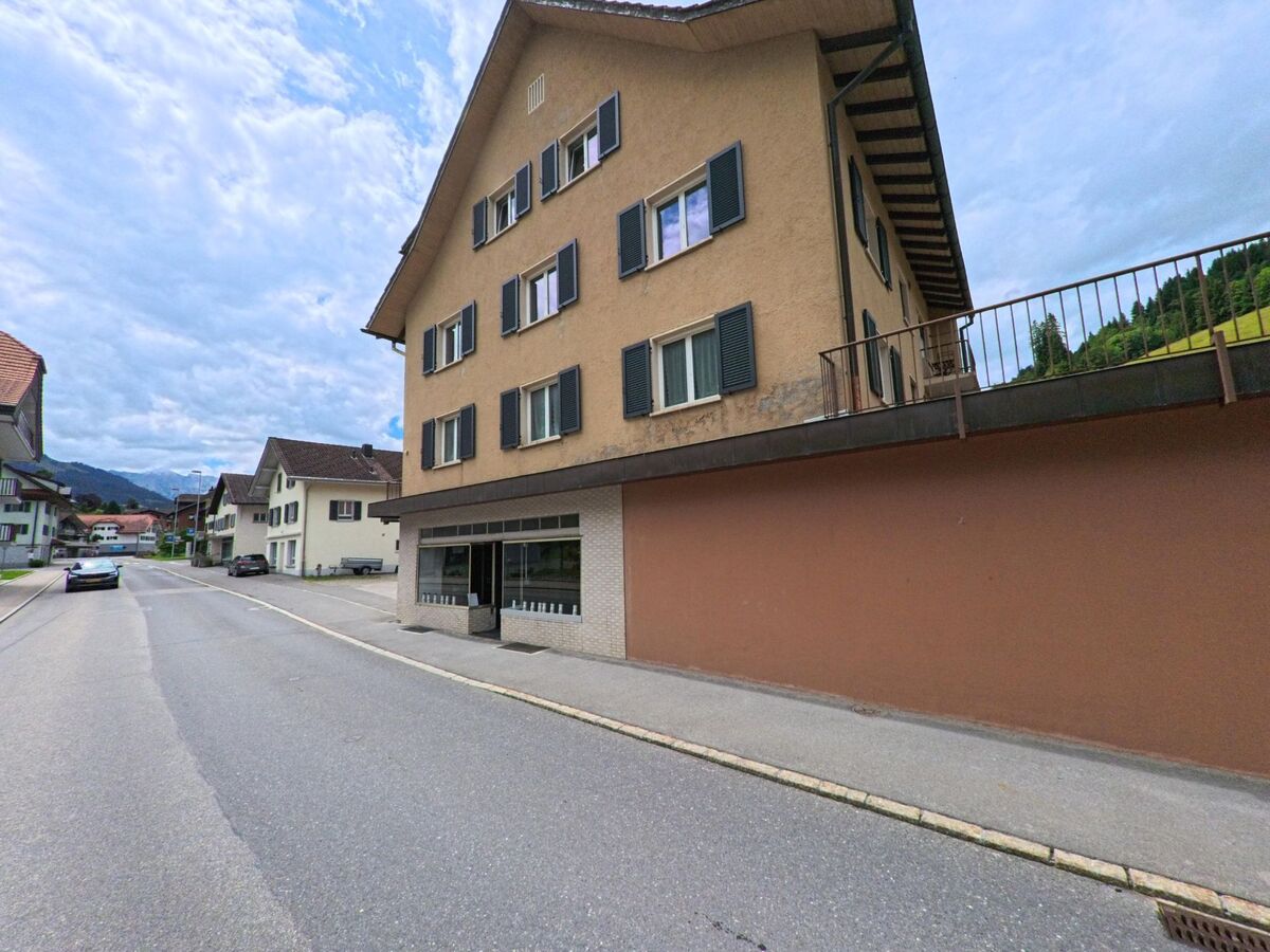 In Flühli (PLZ 6173, Kanton Luzern) steht ein mehrstöckiges Gebäude mit Ladenlokal entlang einer geraden Wohnstrasse am Rande des Dorfzentrums.