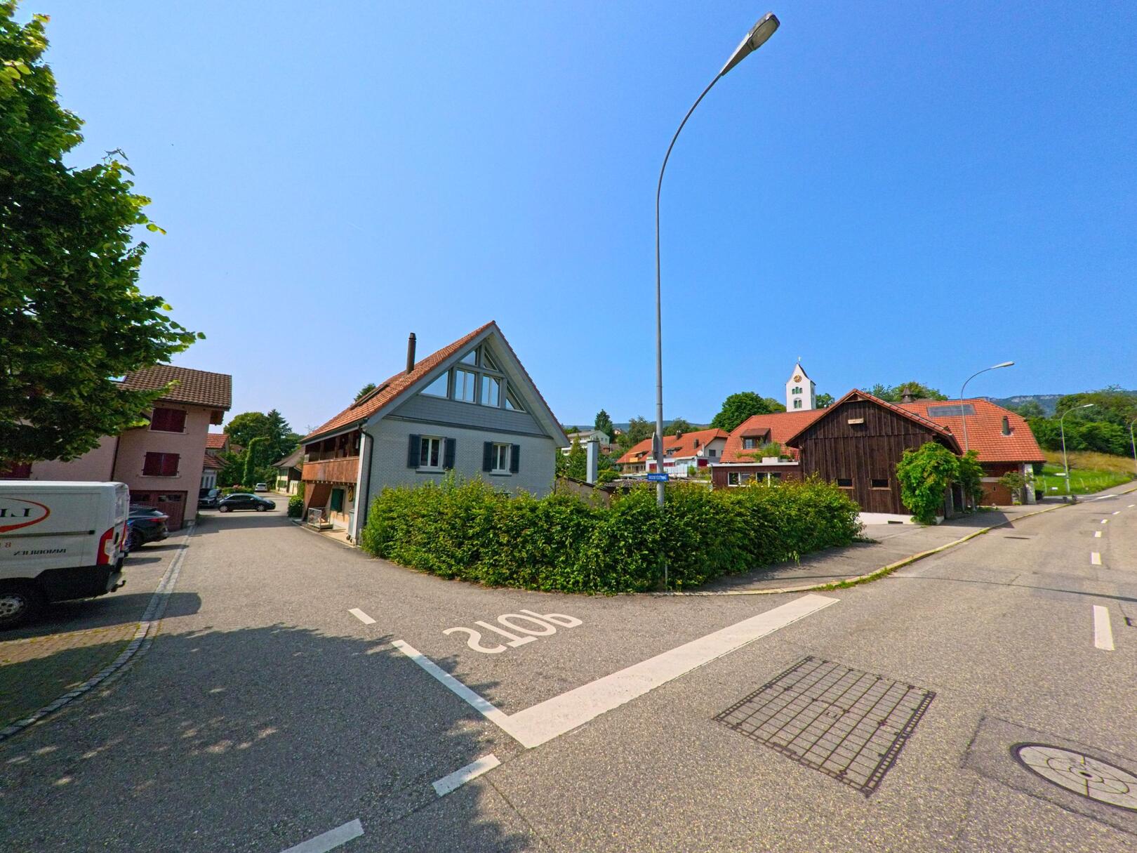 In Flumenthal (4534, Kanton Solothurn) gruppieren sich Wohnhäuser und ein dunkles Scheunengebäude entlang einer ruhigen Strasse mit Blick auf den Kirchturm.