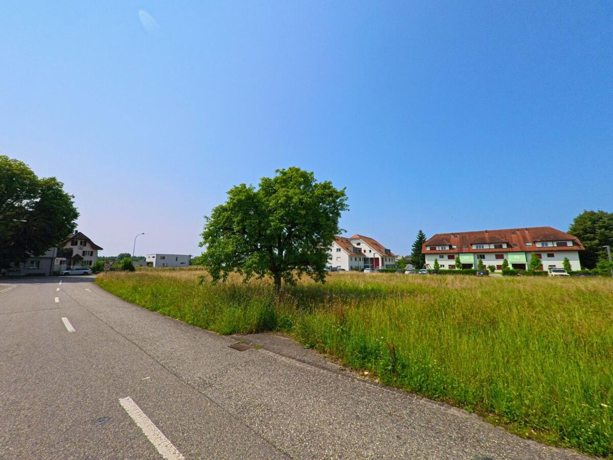 Zwischen einer Landstrasse und einer Wiese stehen in Flumenthal (4534, Kanton Solothurn) mehrere Mehrfamilienhäuser mit roten Dächern und grünen Fensterläden.