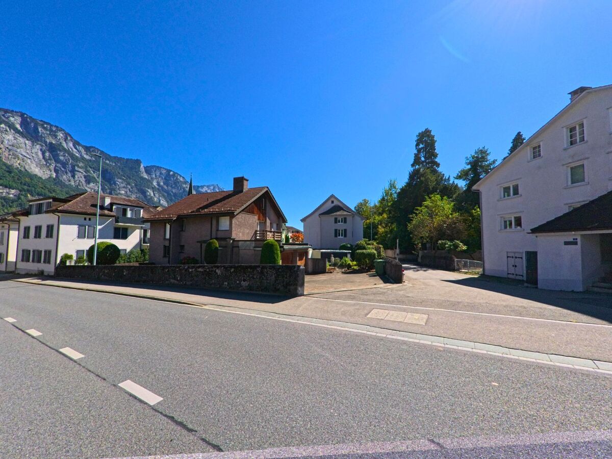 In Flums (8890, Kanton St. Gallen) stehen Einfamilienhaeuser entlang einer breiten Strasse mit Blick auf die steilen Felswaende der umliegenden Berge.
