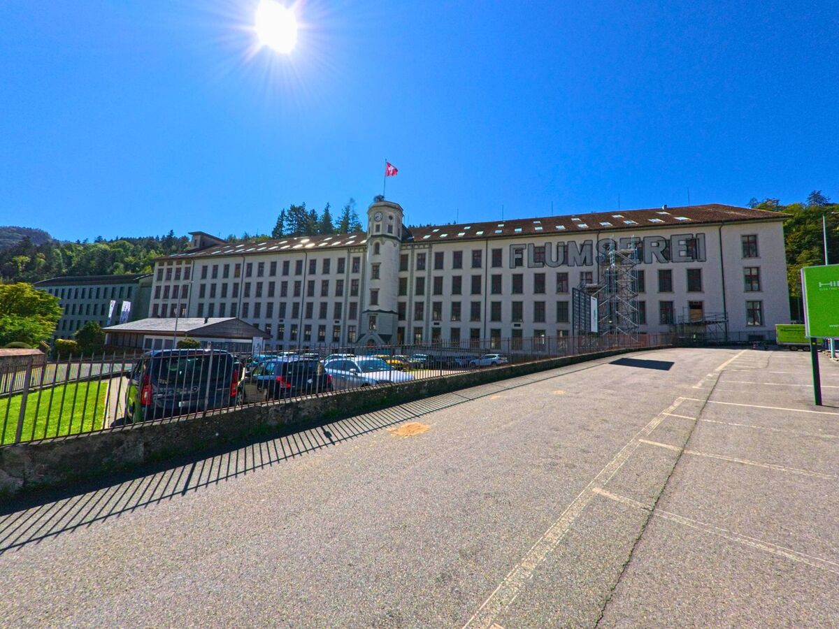 Das markante Gebaeude der ehemaligen Textilfabrik Flumserei in Flums (8890, Kanton St. Gallen) steht unter blauem Himmel mit parkenden Autos im Vordergrund.