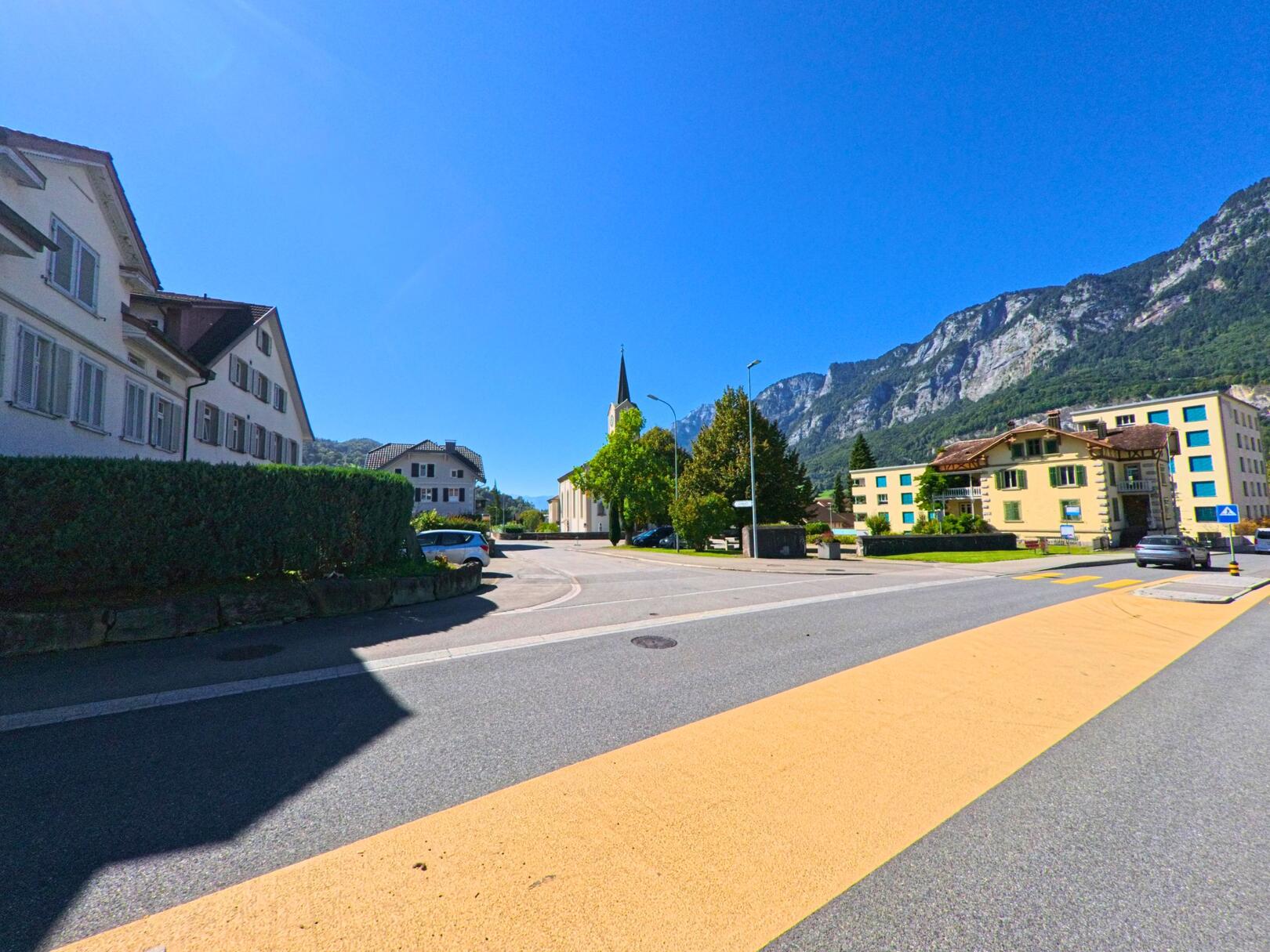Im Zentrum von Flums (8890, Kanton St. Gallen) fuehrt eine Strasse mit gelbem Fussgaengerstreifen an Wohnhaeusern und der Kirche vorbei, eingerahmt von markanten Felswaenden.