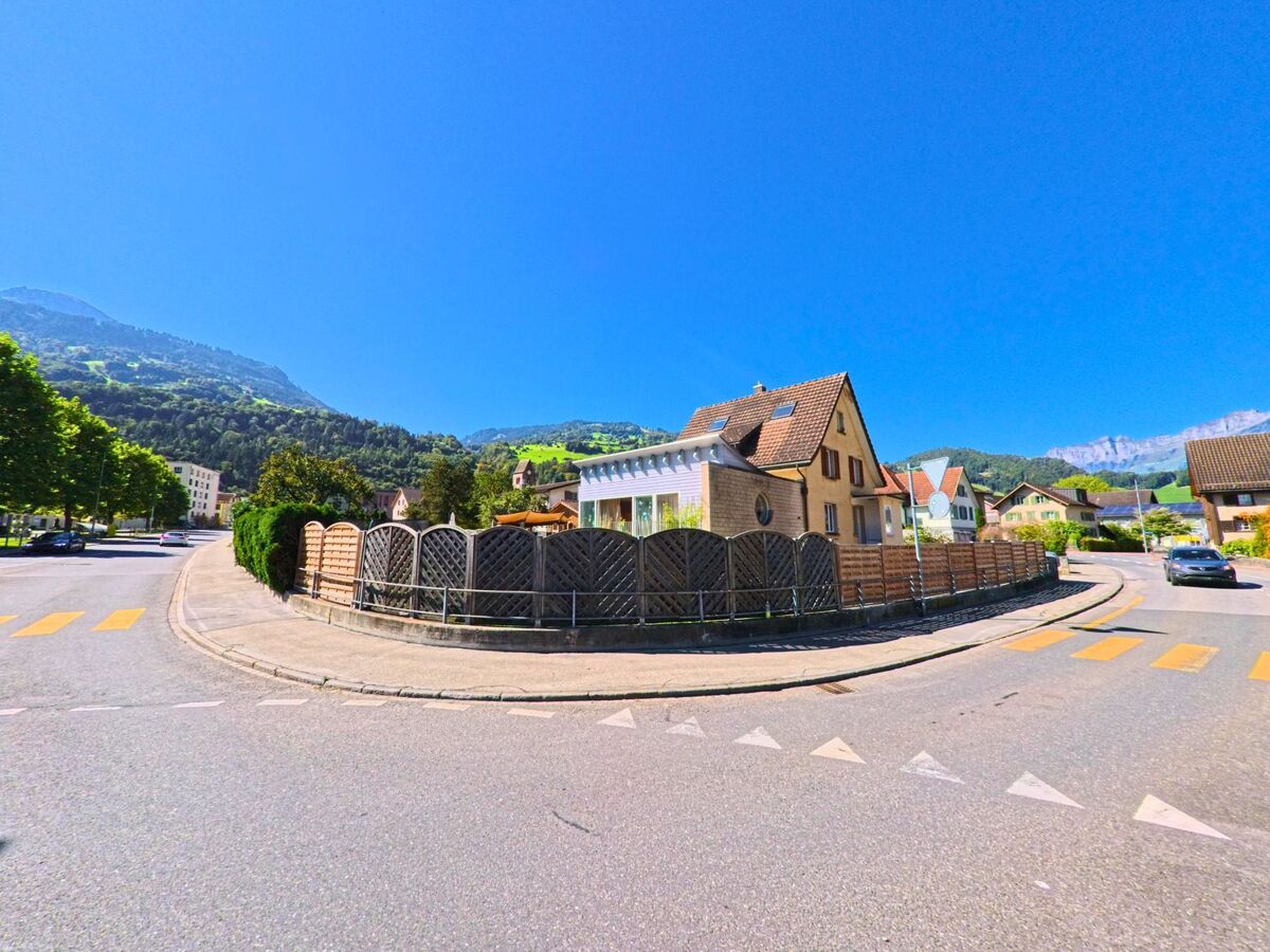 An einer Strassenkreuzung in Flums (8890, Kanton St. Gallen) stehen Einfamilienhaeuser mit Holzzaeunen vor gruenen Haengen und klarer Bergkulisse.