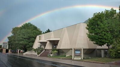 Vor dem modernen Gebäude der Fondation Pierre Gianadda in Martigny (1920, Kanton Wallis) spannt sich ein Regenbogen über den Himmel, während die nasse Strasse im Vordergrund glänzt.