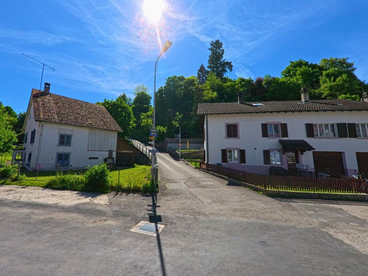 Am Fuss eines bewaldeten Hangs in Fontenais (2902 im Kanton Jura) stehen zwei Wohnhäuser beidseits einer ansteigenden Strasse im Gegenlicht der Sonne.