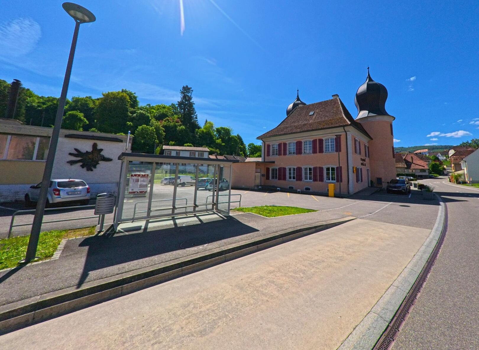 Vor dem markanten Gebäude mit Zwiebeltürmen in Fontenais (2902 im Kanton Jura) liegt eine überdachte Bushaltestelle mit Blick auf die angrenzende Hauptstrasse.