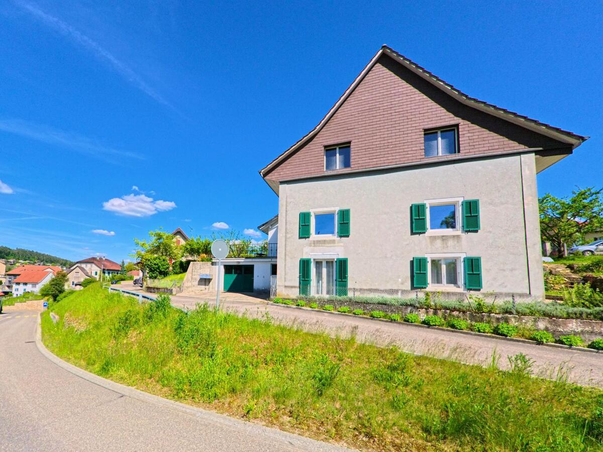 In Fontenais (2902 im Kanton Jura) steht ein Wohnhaus mit grünen Fensterläden an einer sanft geschwungenen Hangstrasse mit Blick auf das Quartier.
