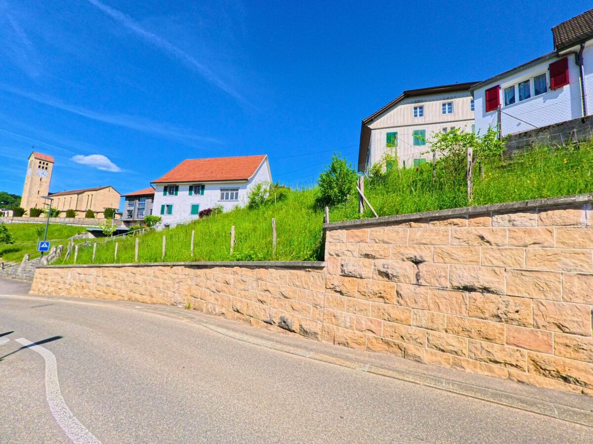 Oberhalb einer Stützmauer in Fontenais (2902 im Kanton Jura) stehen mehrere Wohnhäuser mit Blick auf die markante Kirche mit Uhrturm im Hintergrund.