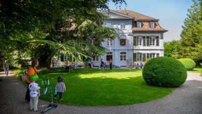 Im Forum Schlossplatz in Aarau (5000, Kanton Aargau) geniessen Besucher den sonnigen Garten vor dem historischen Gebaeude mit gepflegtem Rasen und grossen Baeumen.