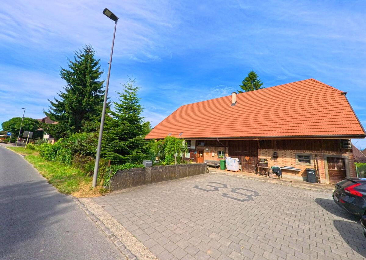 In Fräschels (3284, Kanton Freiburg) liegt ein traditionelles Bauernhaus mit rotem Ziegeldach direkt an einer gepflasterten Einfahrt, umgeben von Tannen und Sträuchern.