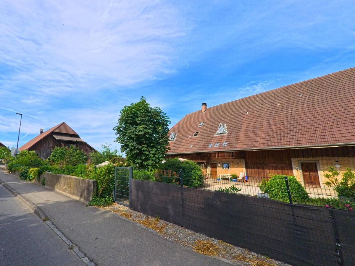In Fräschels (3284, Kanton Freiburg) steht ein breites Bauernhaus mit rotem Ziegeldach und gepflegtem Vorgarten entlang einer ruhigen Strasse.