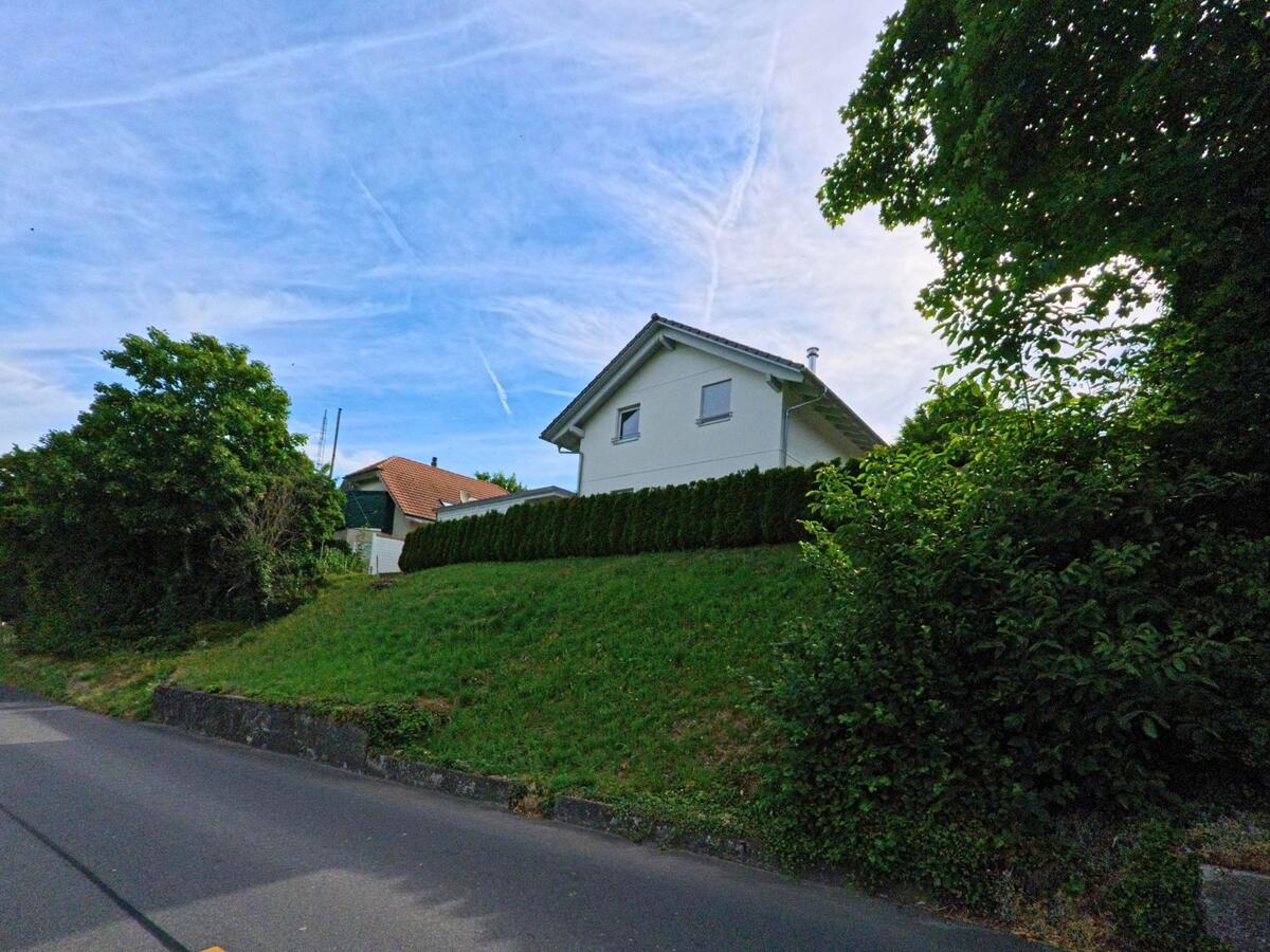 Ein modernes Einfamilienhaus in Fräschels (3284, Kanton Bern) steht leicht erhöht am Hang, eingerahmt von dichter Vegetation und einer ruhigen Strasse.