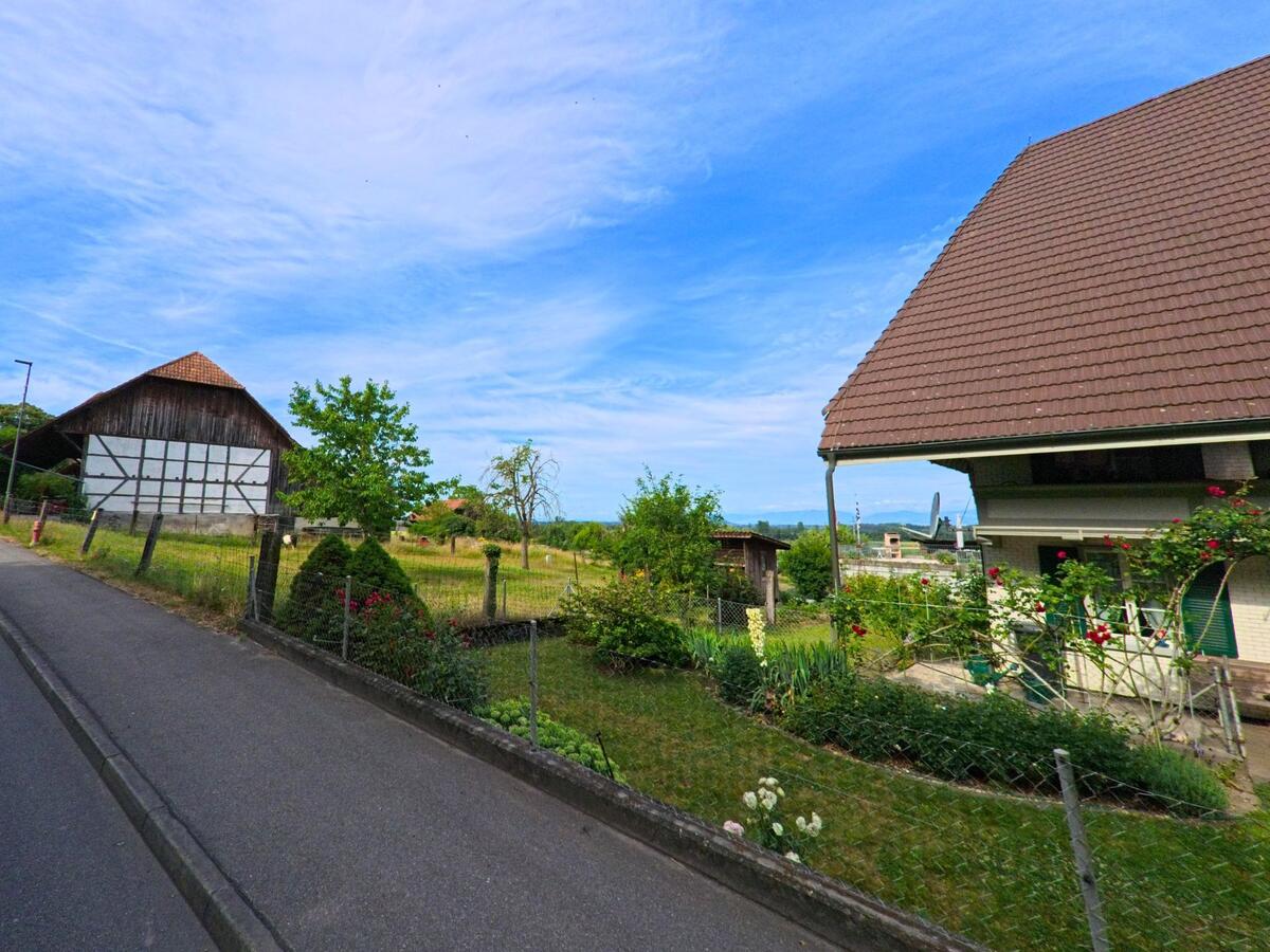 In Fräschels (3284, Kanton Freiburg) erstreckt sich entlang einer schmalen Strasse ein Gehöft mit Fachwerkscheune, Garten und Blick auf die weite Landschaft.