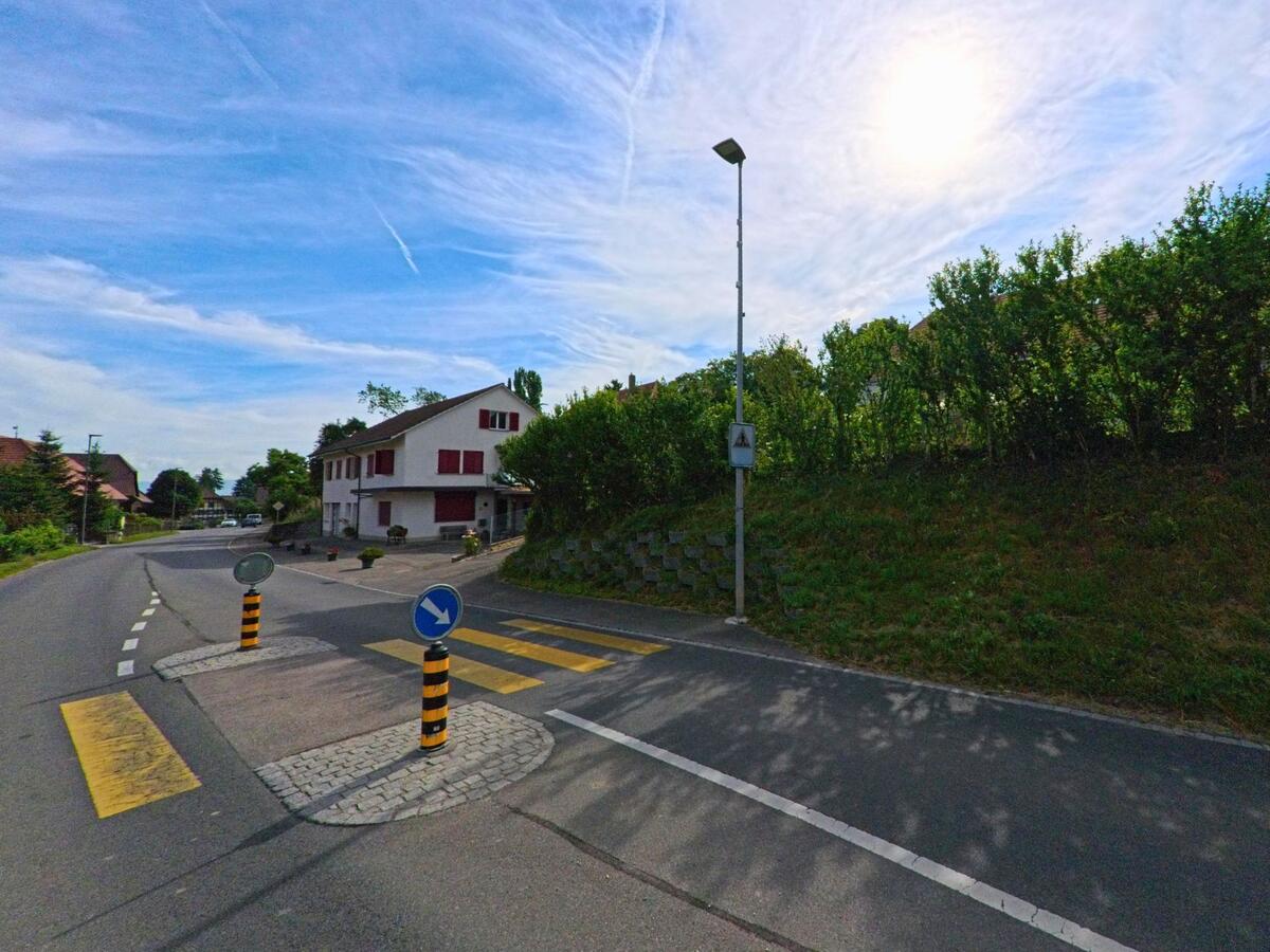 Eine Verkehrsinsel mit Fussgängerstreifen markiert in Fräschels (3284, Kanton Bern) den Übergang zu einer Wohnstrasse mit typischem Dorfhaus und roten Fensterläden.