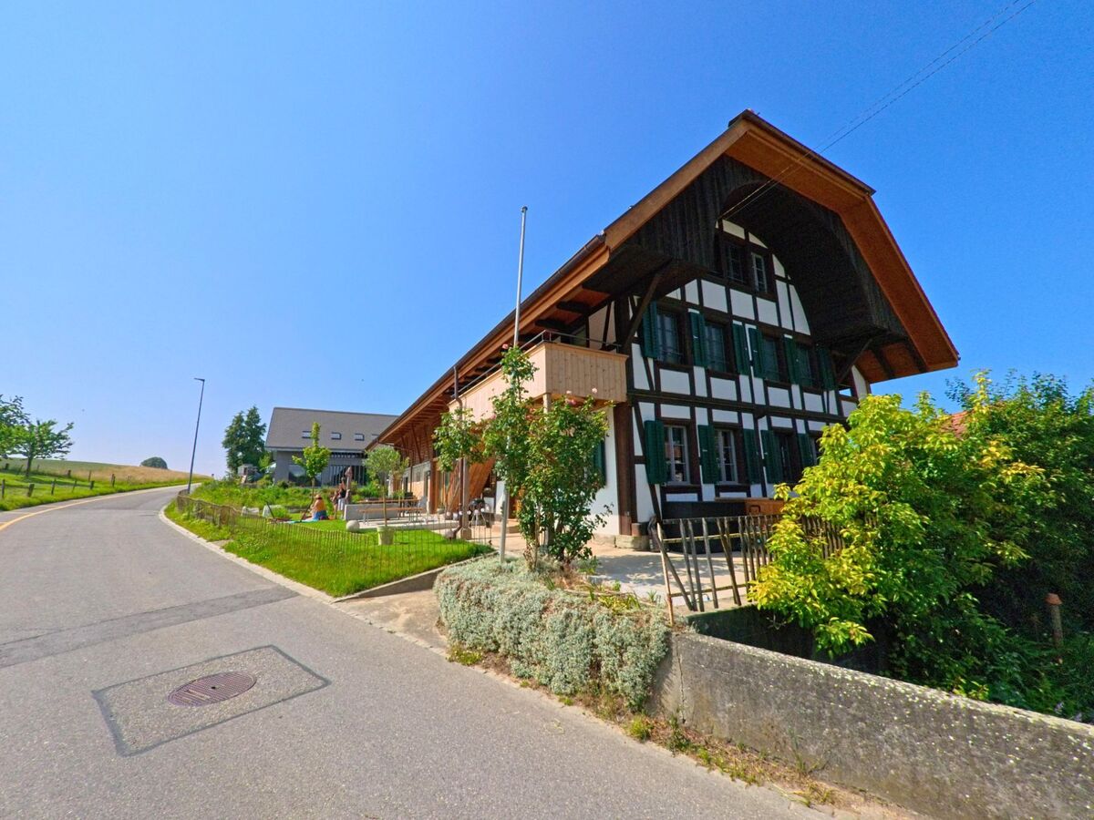 In Fraubrunnen (3313, Kanton Bern) führt eine Straße an einem traditionellen Fachwerkhaus mit grünen Fensterläden und einem gepflegten Garten vorbei, während sanfte Hügel im Hintergrund zu sehen sind.