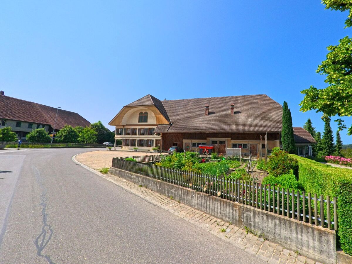 In Fraubrunnen (3313, Kanton Bern) führt eine kurvige Straße an einem traditionellen Gebäude vorbei, das von gepflegten Gärten und grünen Hecken gesäumt ist.