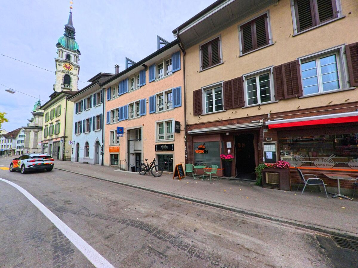 Vor farbigen Altstadthäusern mit Fensterläden und einem Kirchturm im Hintergrund verläuft eine Strasse in Frauenfeld (8500, Kanton Thurgau).