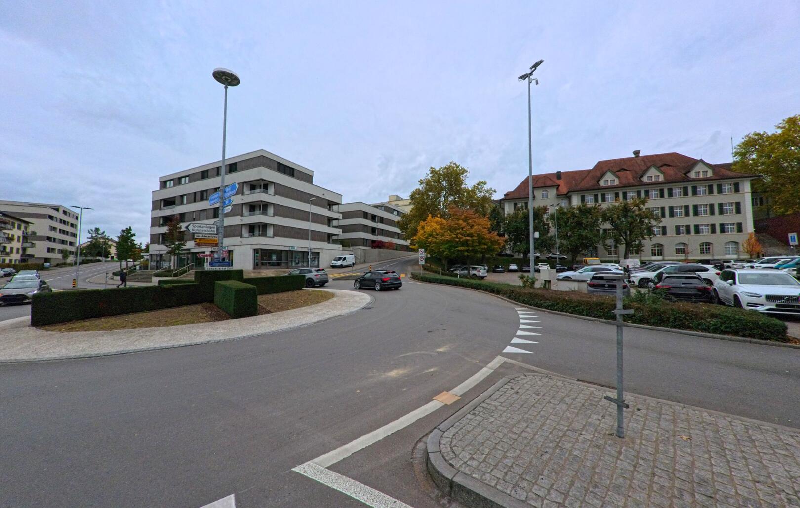 An einer breiten Strassenkurve in Frauenfeld (8500, Kanton Thurgau) liegen moderne Verwaltungsgebäude, ein Parkplatz und herbstlich gefärbte Bäume unter bewölktem Himmel.