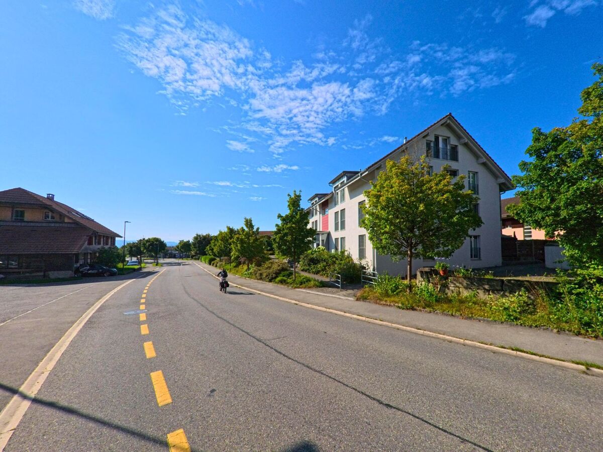Eine breite Strasse in Frauenkappelen (3202, Kanton Bern) fuehrt zwischen Wohngebaeuden und kleinen Baeumen leicht bergab.