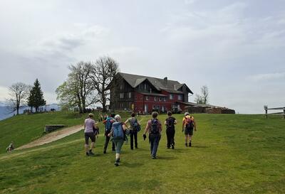 Die Mitglieder des Frauenturnvereins Isenthal aus Isenthal (6461, Kanton Uri) wandern gemeinsam über eine grüne Bergwiese zu einem traditionellen Gebäude in alpiner Umgebung.