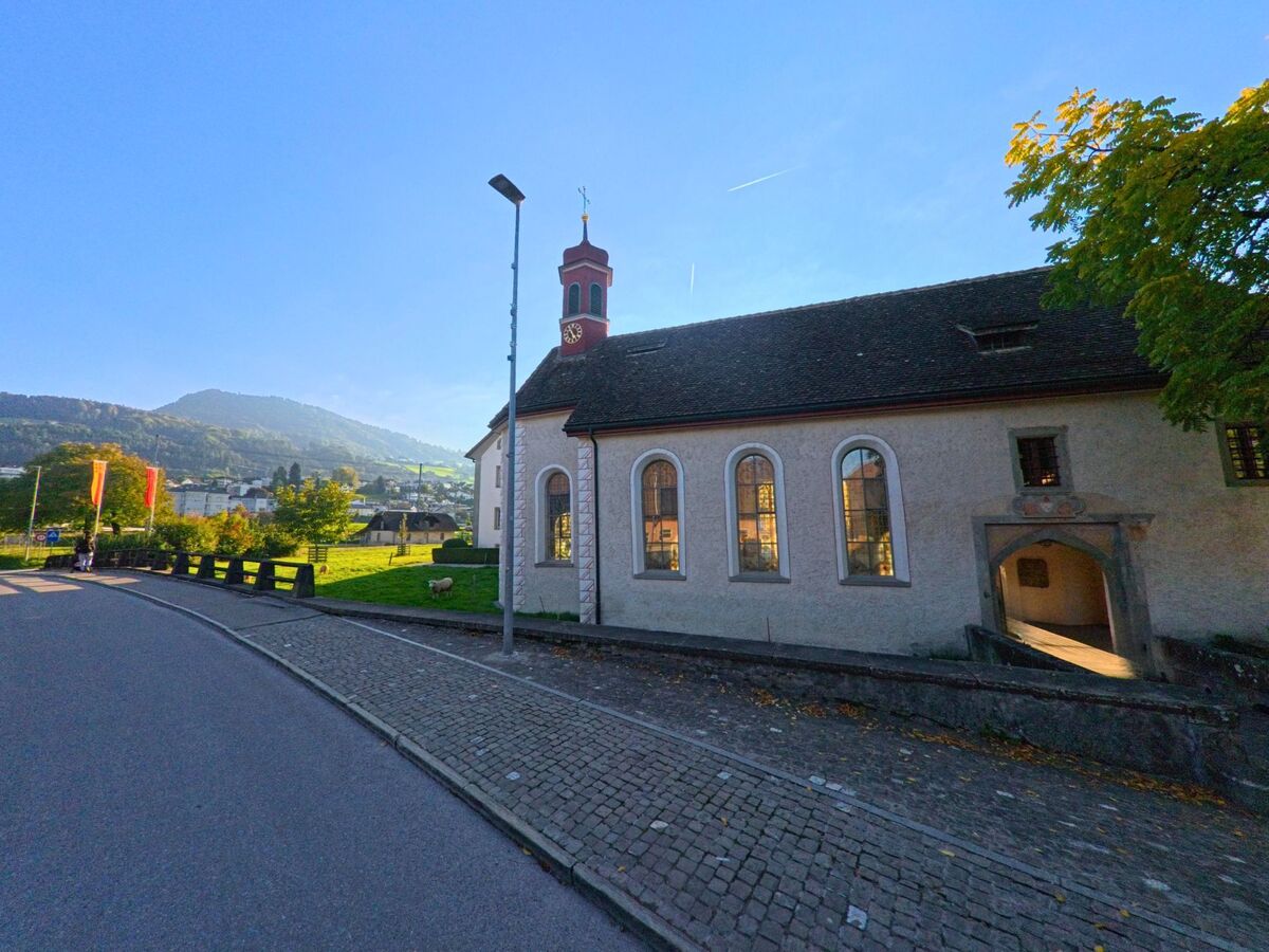 Eine historische Kapelle in Freienbach (8808, Kanton Schwyz) steht am Rand eines sanft geschwungenen Weges, waehrend dahinter gruene Wiesen und Huegel aufragen.