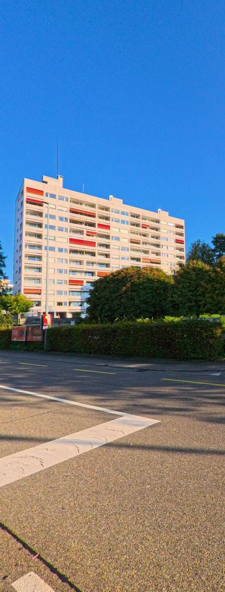 Vor dem hohen Wohngebaeude in Freienbach (8808, Kanton Schwyz) liegt eine ruhige Strasse, waehrend dichtes Gruen den unteren Fassadenbereich teilweise verdeckt.