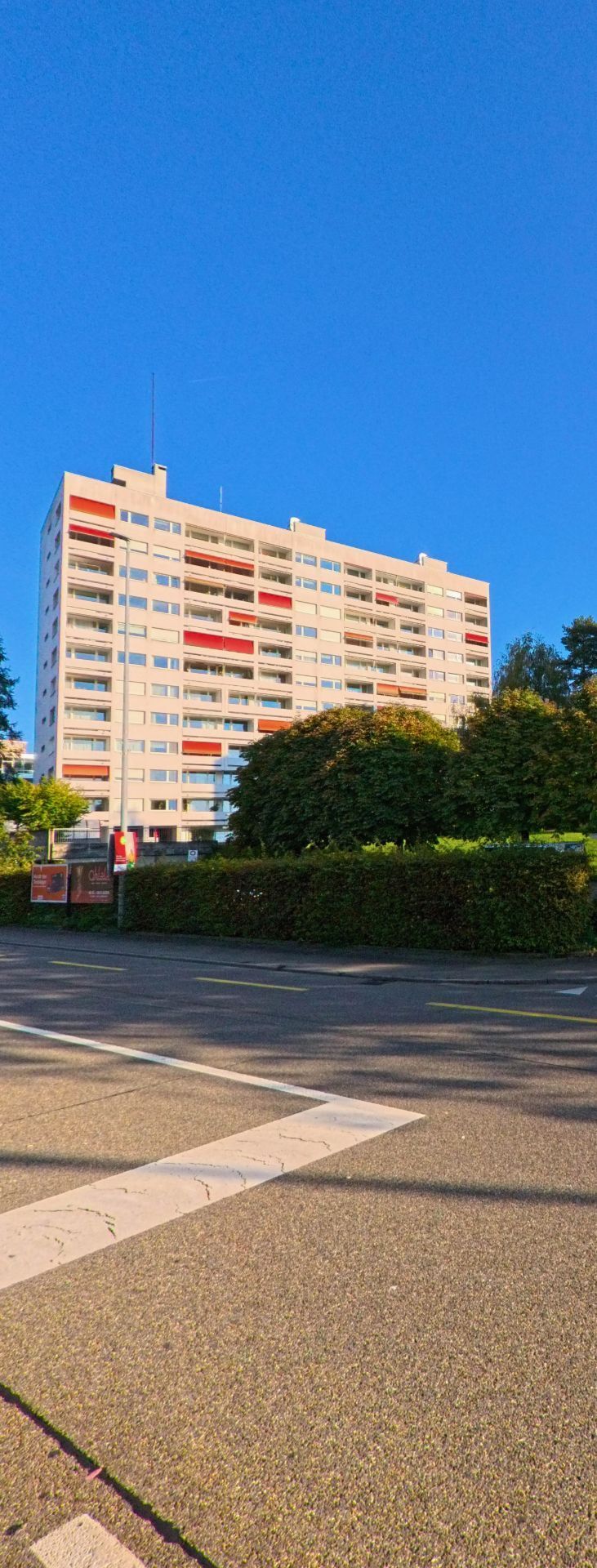 Vor dem hohen Wohngebaeude in Freienbach (8808, Kanton Schwyz) liegt eine ruhige Strasse, waehrend dichtes Gruen den unteren Fassadenbereich teilweise verdeckt.