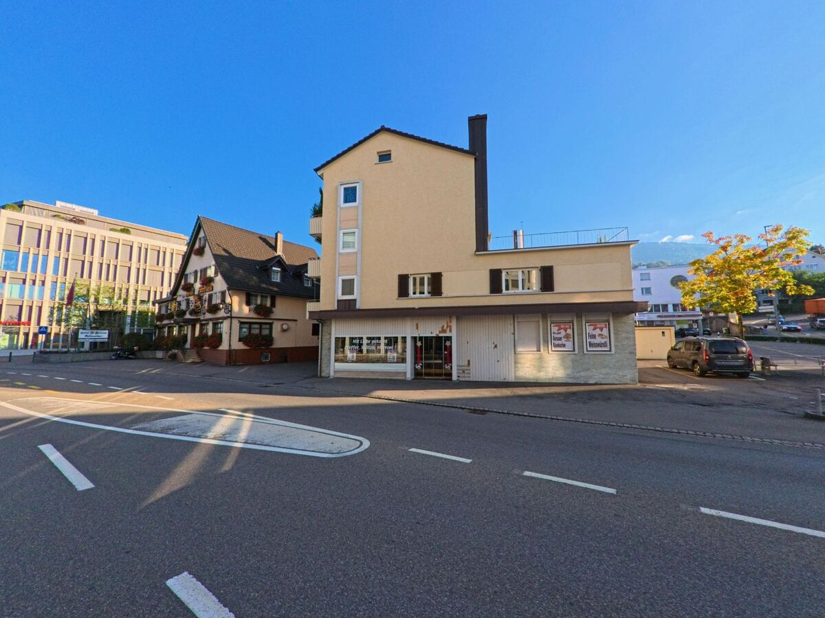 Neben einem traditionellen Haus mit Blumenschmuck steht in Freienbach (8808, Kanton Schwyz) ein schlichtes Gebaeude an einer breiten Strasse im warmen Abendlicht.