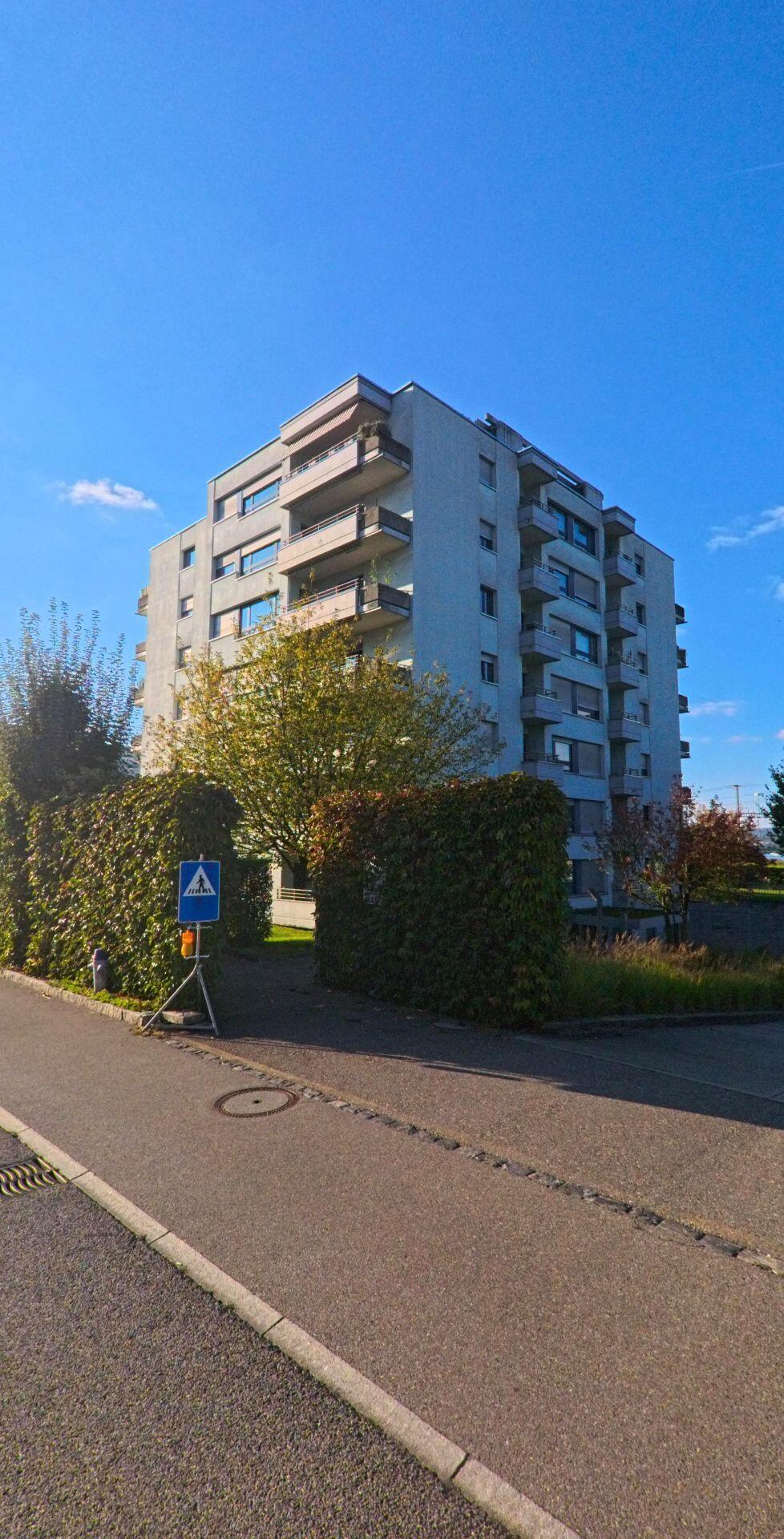 Ein mehrgeschossiges Wohngebaeude in Freienbach (8808, Kanton Schwyz) erhebt sich hinter dichten Hecken, waehrend ein Fussgaengerschild den ruhigen Weg markiert.