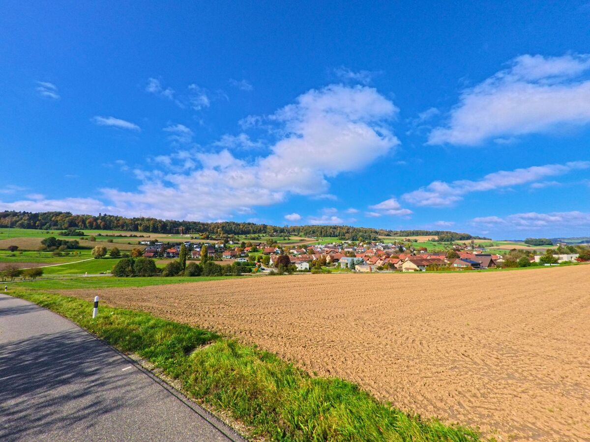 Über einem weiten Acker eröffnet sich der Blick auf das Dorf Freienwil (5423, Kanton Aargau), umgeben von grünen Hügeln und blauem Himmel.